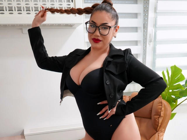 EllaSweety with big tits, black jacket, and glasses, holding her brown braid, wearing a black bodysuit, standing in a room with a tan chair and green plant.