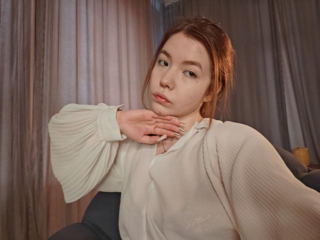 NidaSaltsman is a young woman with fair skin and red hair in a loose updo. She wears a white ribbed blouse with puffed sleeves and grey high-waist pants. Her right hand is gently touching her neck. She has a small beauty mark under her right eye. The background includes grey curtains. The lighting is soft, highlighting her calm expression. Her nails are long and manicured. The image has a casual, intimate feel.