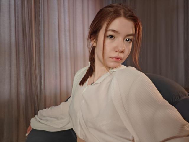 NidaSaltsman, a young woman with fair skin and brown hair in two pigtails, sits in a dark blue chair. She wears a loose, white, long-sleeve shirt. Her expression is neutral, slightly pouty. The image has soft lighting, with warm tones. Her hair is slightly messy, and her eyes are focused on the camera. The background is a blurred curtain. The composition is centered, with NidaSaltsman slightly turned to her right. The texture of her shirt is smooth and slightly reflective.