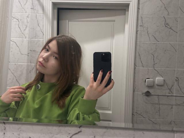 NidaSaltsman takes a selfie in a bathroom mirror. She has light brown hair, fair skin, and a neutral expression. She wears a bright green, button-up shirt with puffed sleeves. Her right hand holds a black smartphone, and her left hand touches her shirt. The marble-tiled wall has visible cracks. The door behind her is white with a simple design. Her nails are painted red. The mirror reflects her upper body and face.
