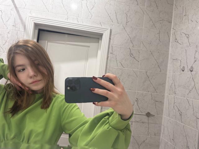A selfie of NidaSaltsman in a bathroom. She has light skin, shoulder-length brown hair, and is wearing a green, long-sleeve, puffy shirt. She holds a green phone with red-painted nails in her right hand, resting her left hand on her head. The marble-tiled wall has a grey, veined pattern. The white door is in the background. Her expression is neutral, slightly bored. The reflection in the mirror is clear.