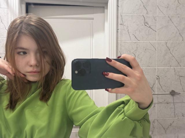 NidaSaltsman with shoulder-length brown hair, green sweater, and dark red nails, taking a selfie in a bathroom with a white door and marble tiles, holding a black phone.