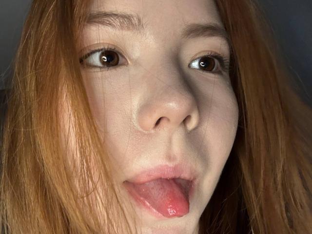 NidaSaltsman's face with red hair, brown eyes, and tongue slightly out, showing her pink tongue and slightly parted lips. Her skin is pale with subtle freckles.