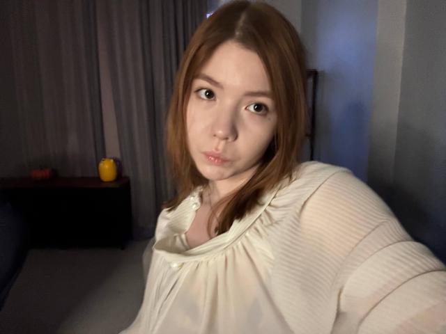 NidaSaltsman with light brown hair, pouty lips, and green eyes, wearing a white ribbed shirt, in a dimly lit room, showing a small, wet, red mark on her chest.