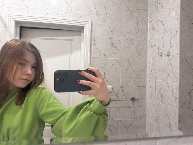 NidaSaltsman, a young woman with light skin and straight brown hair, takes a mirror selfie. She wears a loose, bright green long-sleeve shirt with puffy sleeves. Her right hand, with dark red nail polish, holds a black smartphone. The background features a white door with a marble-patterned tile wall. The mirror reflects her slightly annoyed expression. The composition is simple, focusing on her upper body and the reflection in the mirror. The image has a casual, everyday style.