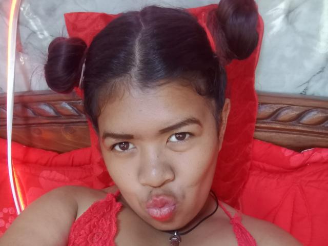 MariahSquirting69 has dark brown skin and wears her black hair in two small buns. She's making a pouty face with her lips slightly pursed. She's wearing a red lace halter top and a black choker with a small charm. Her eyebrows are thick, and she has brown eyes. The red lace and buns contrast with her skin tone. The image has a warm, soft light. The background is blurred but includes a wooden headboard and red pillows. The focus is on MariahSquirting69's face and expression.