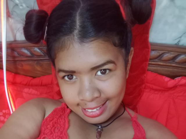 Close-up photo of MariahSquirting69. She has dark brown skin, brown eyes, and a slight smile with pink lipstick. Her hair is in two tight buns on top of her head. She wears a red lace halter top and a black choker with a small pendant. The background is red with a wooden headboard. The lighting is soft, highlighting her face and hair. Her eyebrows are thick, and she has a small beauty mark on her right cheek. The image is sharp with a warm, inviting color palette.