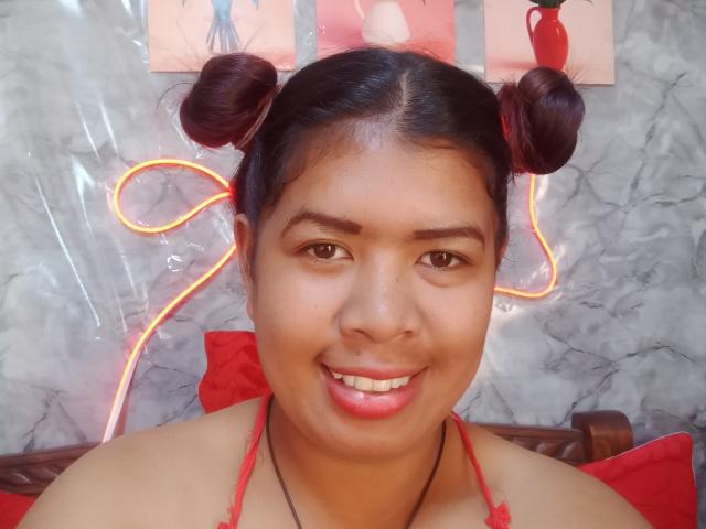 MariahSquirting69 has medium brown skin and dark hair in two buns, with a red string halter top. She smiles with pink lipstick, brown eyes, and a slight overbite. Neon orange light wraps around her hair. Background has marble texture and red pillows. Upper face centered, with red art pieces partially visible. Slightly blurred, natural light. MariahSquirting69's face takes up most of the frame, with a direct, cheerful expression. Image has a casual, real-life feel.