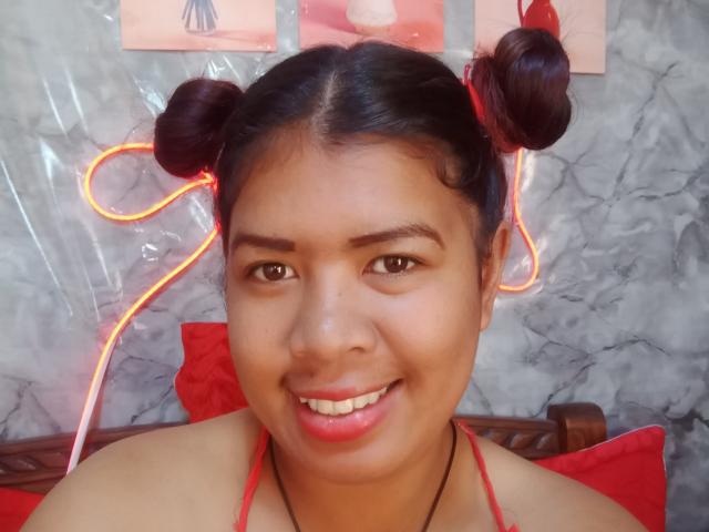 MariahSquirting69 has medium brown skin and dark hair in two small buns, with a bright orange neon line tied in her hair. She's smiling with red lipstick, wearing a red halter top. The background is a marble wall with red and pink posters. The composition is centered on MariahSquirting69's face, with a red cushion and wooden headrest behind her. Her eyes are brown and she has a slight overbite.