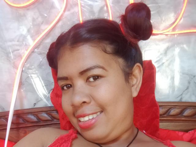 MariahSquirting69 has dark skin, brown eyes, and black double buns. She's smiling with red lips, wearing a red lace top, and neon lights are in the background.