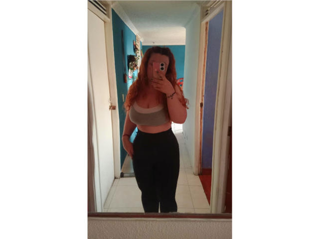 RosseHot6_9 takes a mirror selfie. She has curly, shoulder-length red hair, wearing a gray sports bra and black high-waisted pants. Her right arm is slightly raised, holding a phone with a colorful case. Her left hand rests on her hip. The background includes blue and white walls, a white door on the left, and a doorway on the right. The image is a photograph with a visible reflection in the mirror.