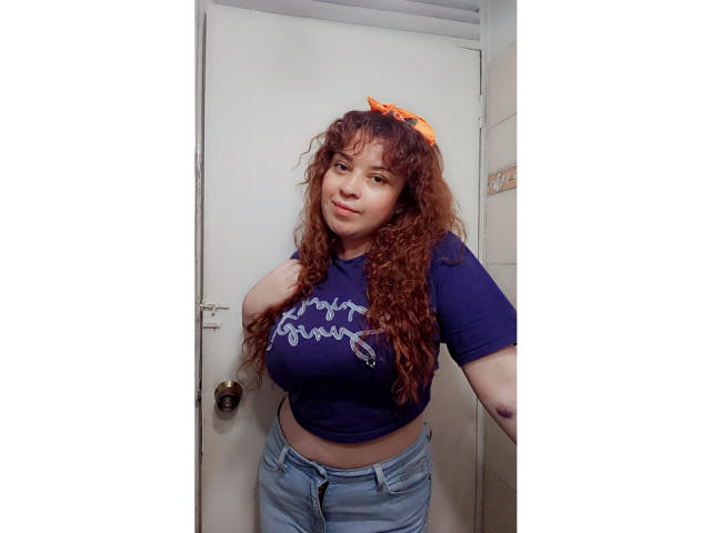 RosseHot69 stands in a doorway, wearing a blue crop top with a white design, and light blue jeans. She has long, curly red hair and a small orange bow on her head. She has a medium build with large breasts. The door behind her is white with a brass doorknob. The image has a casual, everyday style. RosseHot69's expression is neutral, and she has one hand on her hip. The lighting is even, highlighting her features and clothing.