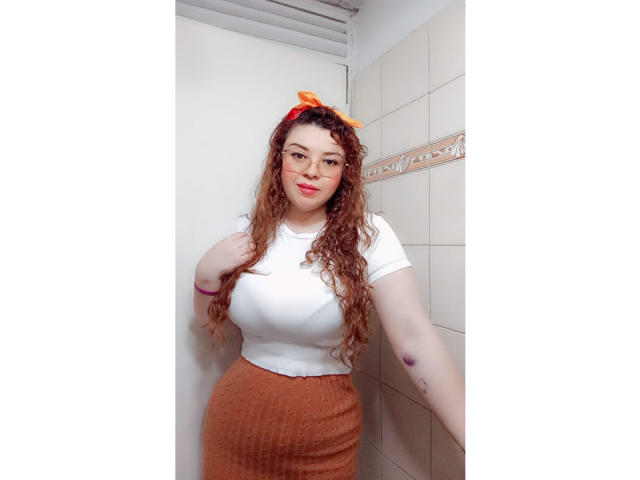 RosseHot69, con i capelli rossi ricci, gli occhiali, la camicia bianca e la gonna marrone, è in piedi in un bagno. Ha il seno grande ed è sorridente, e indossa un fiocco rosso e arancione..
