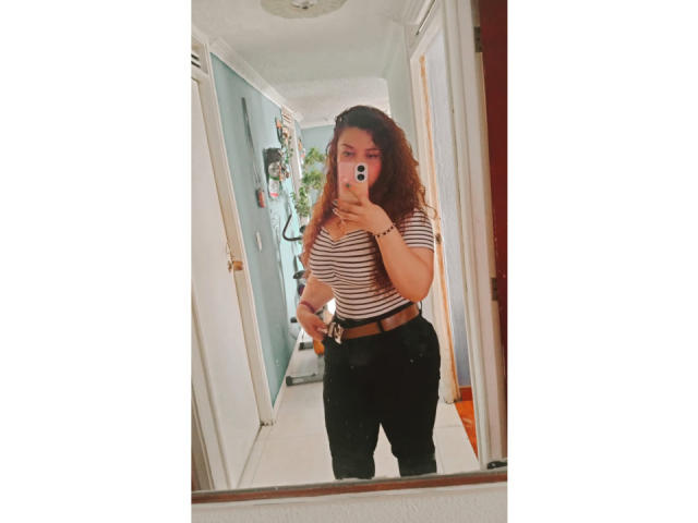 RosseHot6.9 stands in a hallway, taking a mirror selfie. She has curly brown hair and wears a striped, short-sleeved shirt with black pants. Her right hand holds a phone, partially covering her face. She's medium-build with a slightly curvy physique. The mirror reflects a white door and a green plant in the background. The photo has a soft, slightly grainy filter, giving it a vintage look. RosseHot6.9 is centered in the frame.