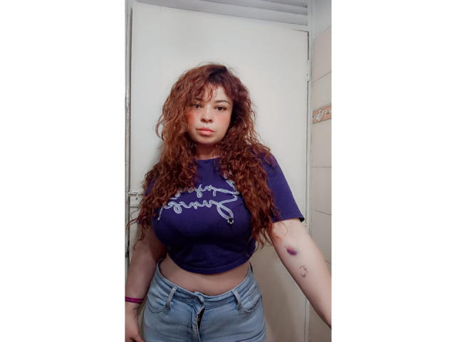 RosseHot69, con i capelli rossi ricci, grandi seni, indossa una camicia blu e jeans, ha un livido sul braccio destro, è in piedi in un bagno e guarda la fotocamera..