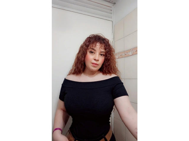 RosseHot69 tiene el pelo rojo rizado, piel clara y lleva una blusa negra con un hombro descubierto, mostrando sus grandes senos, y una pulsera rosa en un baño pequeño..