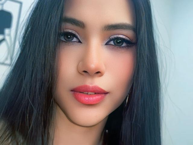 EstrellaXHot's face with long dark hair, pink lips, and purple eyeshadow. She's looking at the camera with a slight smile. Her eyes are dark and she's wearing hoop earrings.