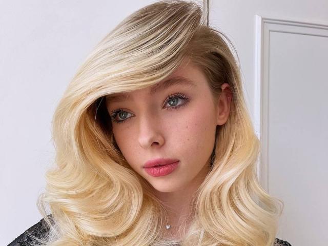 Vivan Wisseman tiene el cabello rubio y ondulado que le cubre un ojo, ojos azules, labios rosados y un rostro ligeramente salpicado de pecas. Lleva una blusa negra..