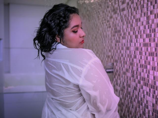 ViickyLee stands in a bathroom with wet, curly black hair and a white, semi-sheer shirt clinging to her back. She looks over her shoulder with red lipstick and closed eyes. The right wall has a mosaic of small, purple-tinted tiles. The left wall is blurred. Her skin is light, and the shirt is slightly transparent, revealing her upper back. The image has a soft, purple-blue light. The overall composition is tight, focusing on ViickyLee's upper body and face.