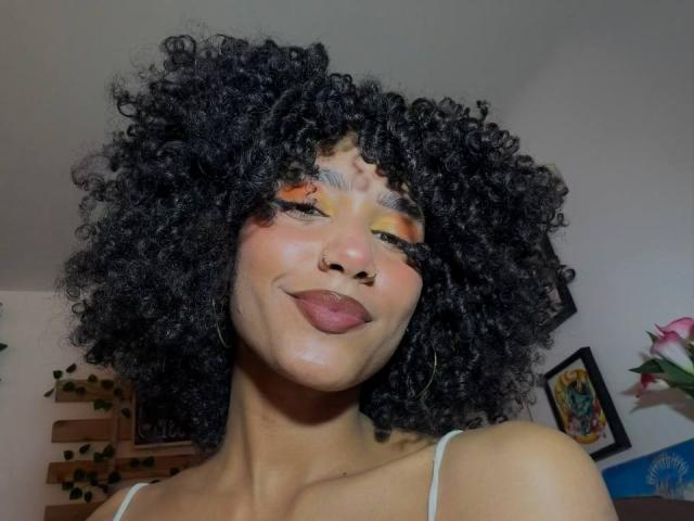 CinthiaWalker with curly black hair, nose ring, and orange eyeshadow, smirking, wearing a white top, showing her smooth brown skin and full lips.