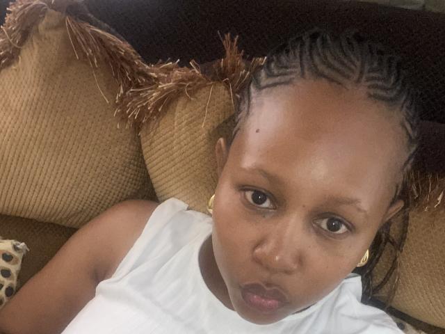 Close-up of SweeOneTee's face, showing a serious expression. SweeOneTee has dark brown skin and is wearing a white sleeveless shirt. She has braided hair with a geometric pattern on top of her head. She's looking directly at the camera with brown eyes. She's wearing small gold earrings. The background includes a brown, textured couch with fringed edges and a patterned pillow. The image is slightly tilted.