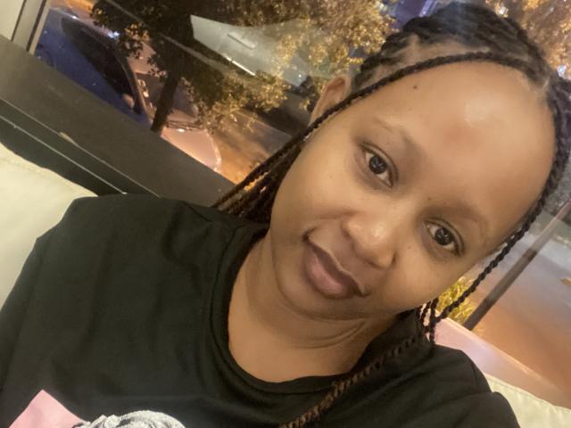 Close-up photo of SweeOneTee, a Black woman with a medium-brown skin tone and braided hair. She's wearing a black t-shirt with a white graphic on the left side. Her lips are painted dark purple. She's sticking out her tongue slightly, giving a playful expression. Her eyes are brown, and she has a small mole on her right cheek. The background is blurry, showing a window with outdoor lights and trees. The lighting is warm.