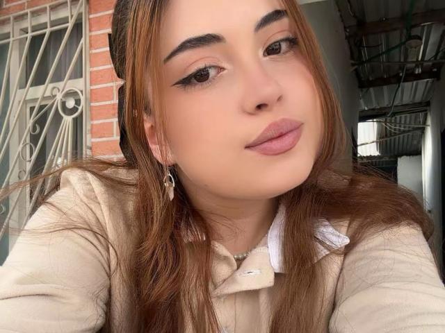 VicluX has light brown skin, long straight brown hair, and wears subtle winged eyeliner with pink lipstick. She has a small nose and full lips. She's wearing a beige top with a white collar. She's looking slightly to the right with a relaxed, confident expression. Small silver hoop earrings are visible. The image is a close-up, focused on her face. The texture of her hair is smooth, and the lighting is natural.