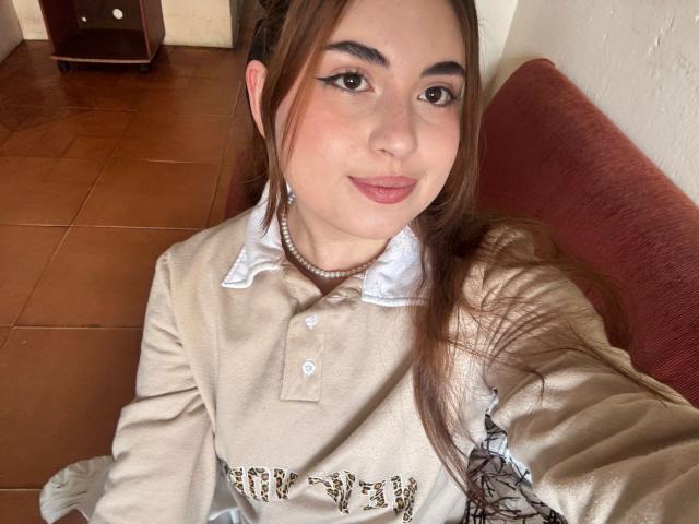 VicluX has light skin, brown eyes, and long brown hair. She's wearing a beige polo shirt with a white collar, a pearl necklace, and has subtle, winged eyeliner. Her lips are painted a soft pink. She's sitting on a red couch, with a brown wooden cabinet and tiled floor in the background. Her hair is slightly messy, and she's smiling gently. The composition is close-up, focusing on her upper body and face. Her relaxed expression and casual outfit create a comfortable, everyday vibe.