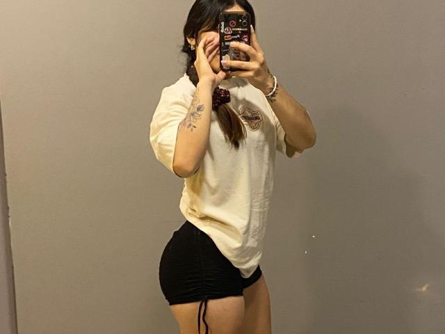 SophiaMarx with braided hair, white tee, and black ruched shorts, holding phone in mirror selfie, showing curvy butt and tattoo on left arm.