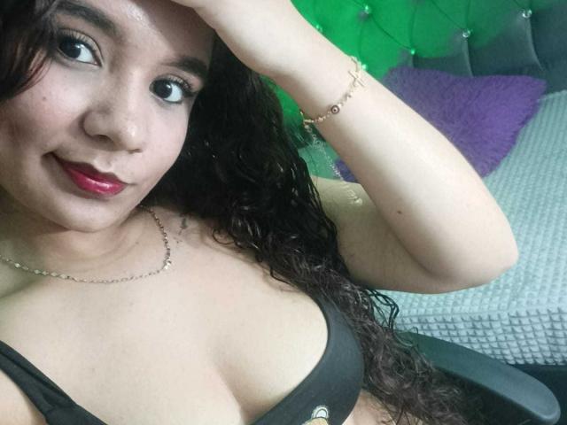 KathyyBlow with curly dark hair, red lipstick, black bra, and bracelet, smiling at the camera, hand raised, showing off her tits, sitting in a chair.