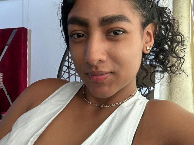 NatalyBlonder with curly black hair, brown eyes, and tan skin, wearing a white sleeveless top, small gold heart earrings, and a thin chain necklace, smiling slightly.