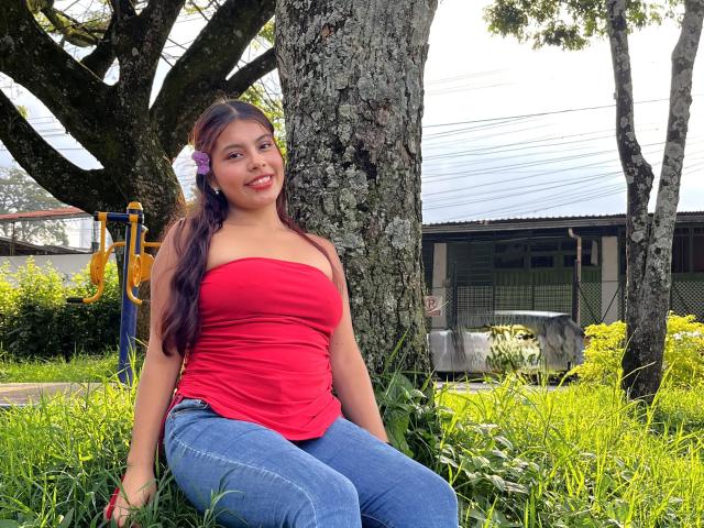 AlyssaGreen sits on grass near a tree, wearing a red strapless top and blue jeans. She has long dark hair with a purple flower on the left. Her red lipstick contrasts with her fair skin. Background includes greenery, a playground structure, and a building with a metal roof. Bright daylight.