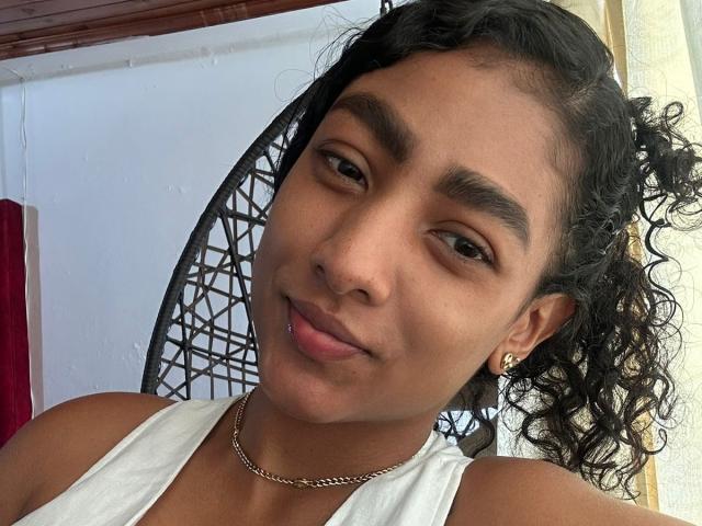NatalyBlonder has medium brown skin, curly black hair in a ponytail, small nose ring, gold earrings, white tank top, subtle smile, and brown eyes, wearing a thin gold chain.