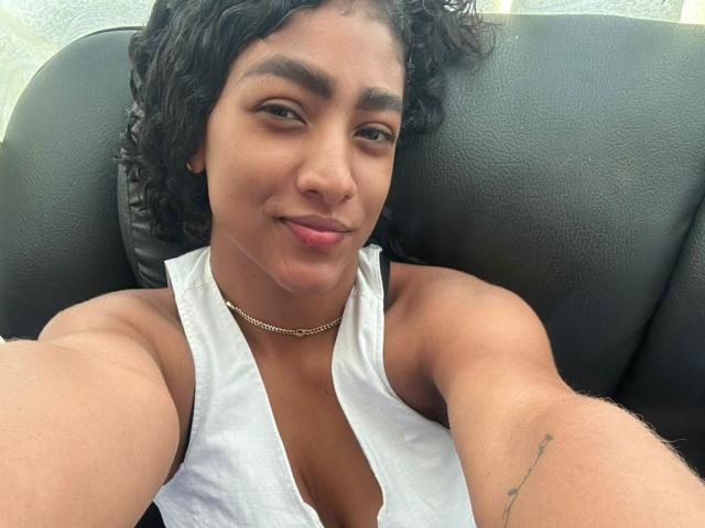 NatalyBlonder with curly black hair, brown skin, and a small tattoo on her right arm, wearing a white halter top and gold chain, smiling at the camera.