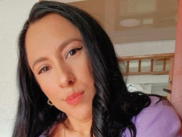 Close-up of JadeFoster with long black hair, light brown skin, and subtle makeup. She has thin black eyeliner and pink lipstick. She's wearing a purple top and a gold necklace. Her expression is calm, with a slight pout. The background is blurred, showing a wooden headboard and part of a ceiling. Natural light highlights her face. Image has a casual, realistic style.