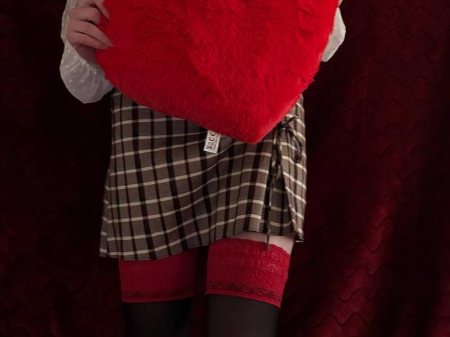 MiyaLia holding a red fuzzy heart, wearing a brown plaid skirt, red lace-topped thigh-highs, and black stockings, with a white shirt partially visible.