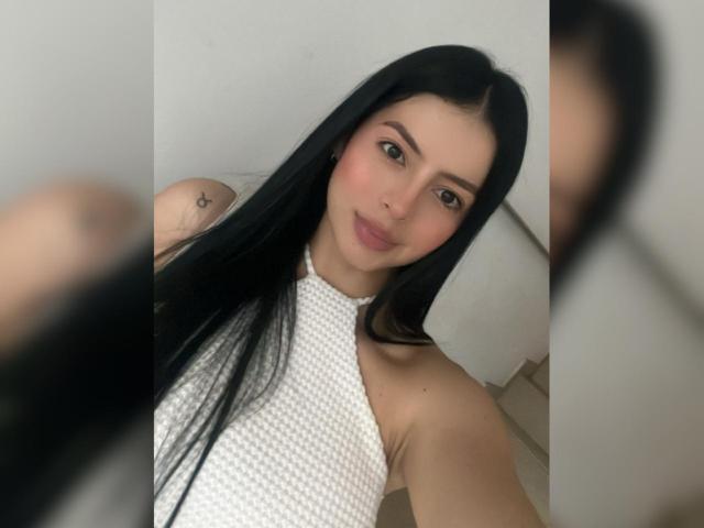 Portrait of IsabellaJames with long black hair, light skin, and brown eyes. She wears a white, textured halter top. Her expression is neutral, with a slight, closed-mouth smile. A small star tattoo is visible on her left shoulder. The background is blurred and minimal. The image is a close-up selfie with a slight blur in the foreground. IsabellaJames's face is centered, and her arm is partially visible. The composition focuses on her upper body and face.