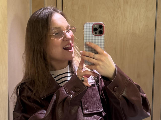 MarikoDentino met een bril, een bruine leren jas, een gestreepte shirt, lachend, met een telefoon met een geruit hoesje in haar hand, haar tong uitgestoken, op een houten achtergrond, met een ring om, die naar haar telefoon kijkt, een spiegel-selfie..
