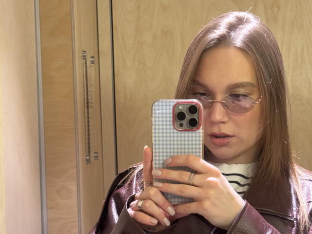Mariko Dentino, con cabello castaño, gafas y un anillo, tomando un selfie con una funda de teléfono a cuadros, vistiendo una chaqueta de cuero marrón y una camisa blanca a rayas..