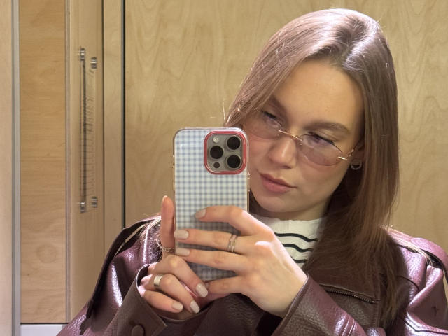 Mariko Dentino, con cabello castaño, gafas y una chaqueta de cuero, se está tomando un selfie con una funda de teléfono a cuadros, mostrando su rostro con los labios fruncidos y su lápiz labial rosa, en un vestuario..