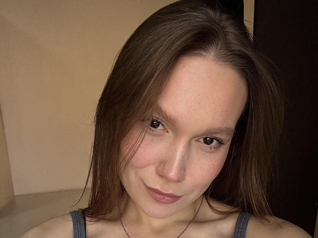 Mariko Dentino mit braunen Haaren, heller Haut und Sommersprossen, die leicht lächelt, trägt ein graues Tanktop und eine feine Halskette, in einem schwach beleuchteten Raum..