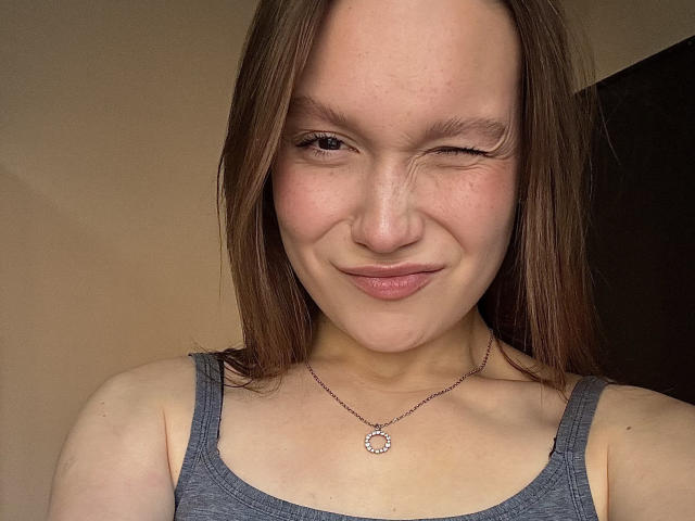 Mariko Dentino ha la pelle chiara, i capelli castani, e sta ammiccando con un piccolo sorriso, indossando una canotta grigia e una collana con un ciondolo circolare..