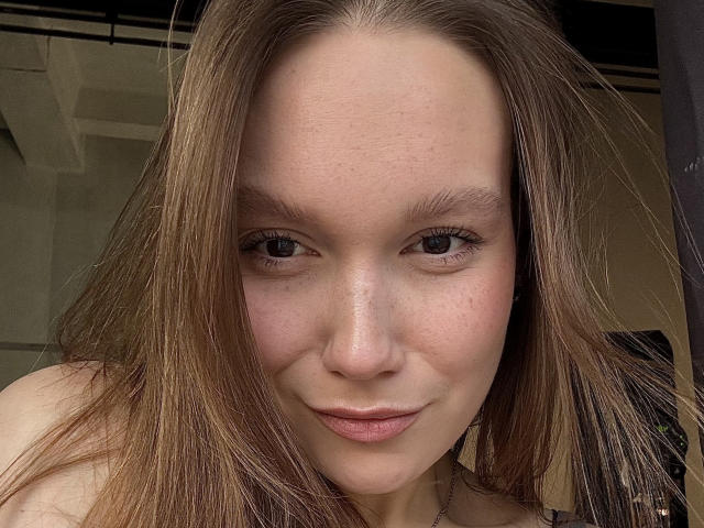 MarikoDentino's face, showing light brown hair, blue eyes, and a slight smile. She has freckles and a subtle pinkish nose, with a small, natural-looking ass in the background.
