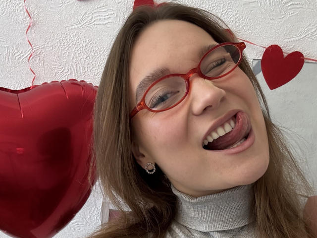 Mariko Dentino, avec des lunettes rouges, la langue tirée, portant un pull gris à col roulé, a un visage mignon, de petites boucles d'oreilles en forme de cœur et des cheveux bruns. Des ballons en forme de cœur rouges sont visibles en arrière-plan..
