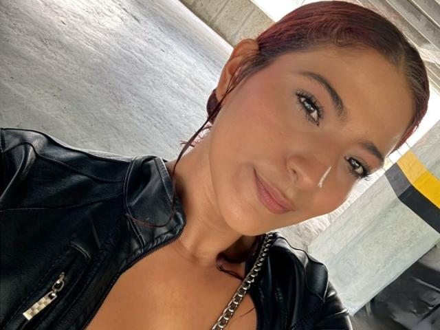 MiaBianchi with red hair, black leather jacket, and chain necklace, smiling at the camera, showing her cute face and small tits. She has a light skin tone and eyeliner.