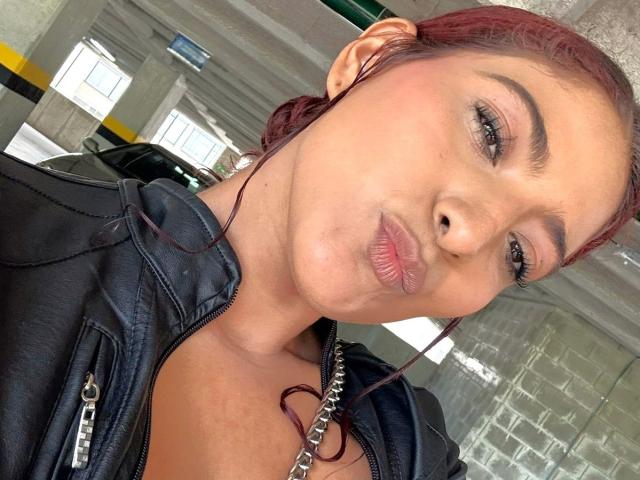 MiaBianchi with puffy pink lips, black leather jacket, red hair, and earbuds, looking at the camera with a slight pout, wearing a chain necklace.