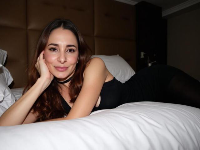 MiaTentacion lies on a bed, head resting on her hand, smiling. She has long, wavy brown hair, light skin, and wears a black choker and a sleeveless black dress. The bed has white pillows and a beige tufted headboard. She's looking directly at the camera, her right cheek slightly raised. The room is dimly lit, with a dark wall in the background. The composition focuses on MiaTentacion's relaxed, confident pose.