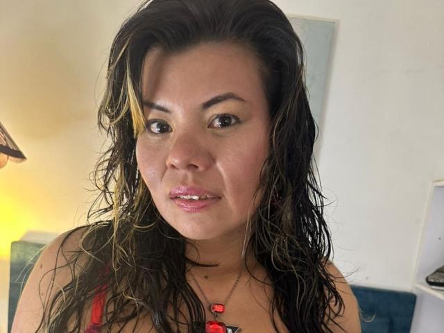 KiaraXMoon tiene el cabello castaño oscuro y húmedo, con reflejos rubios, una cara linda y unos labios rosados y brillantes. Lleva una blusa roja y un collar con cuentas rojas..