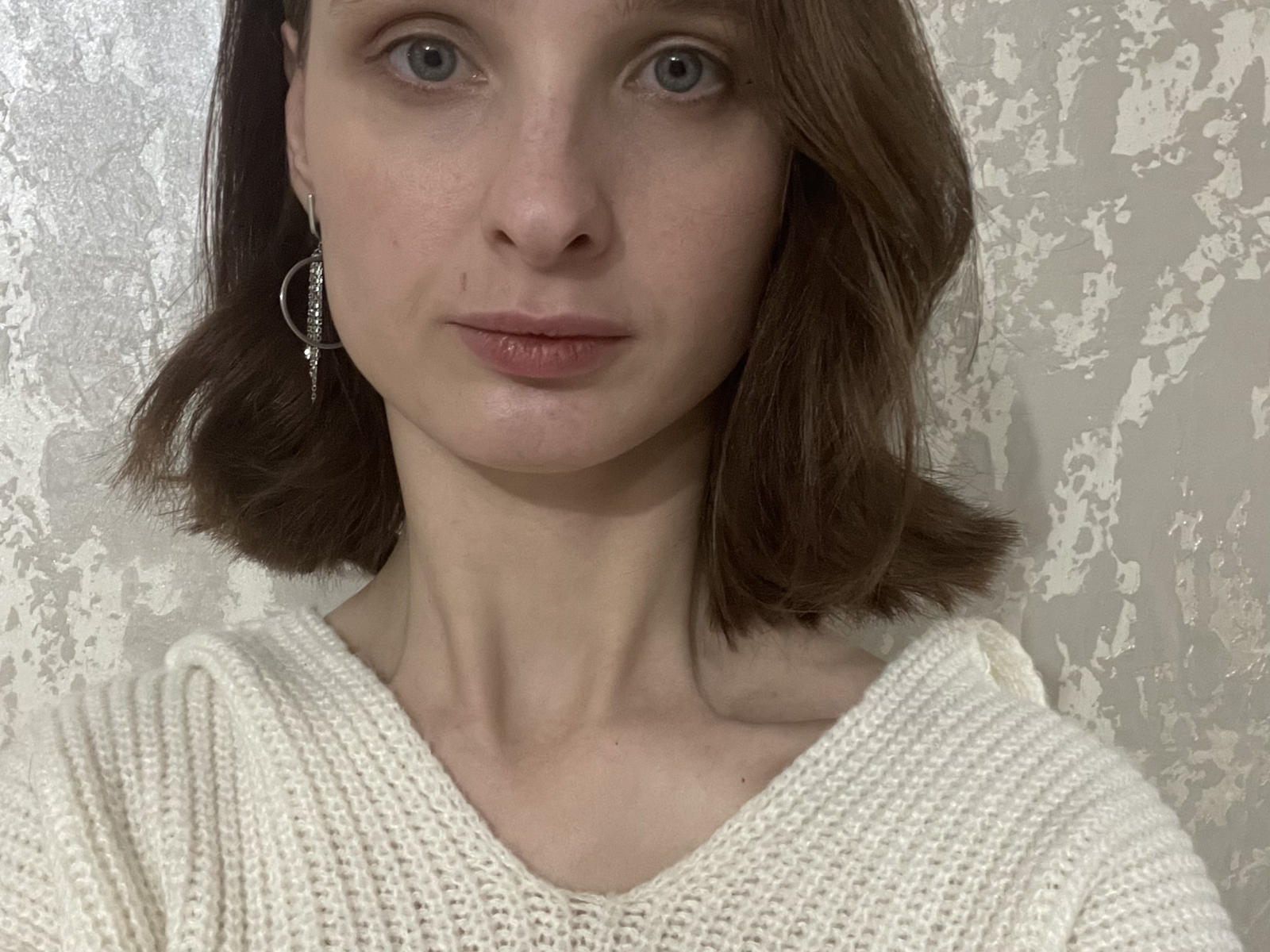 KelsiSchoenthal has fair skin, brown eyes, and brown shoulder-length hair. She's wearing a white knit sweater with a V-neck, and silver hoop earrings. Her face is neutral.