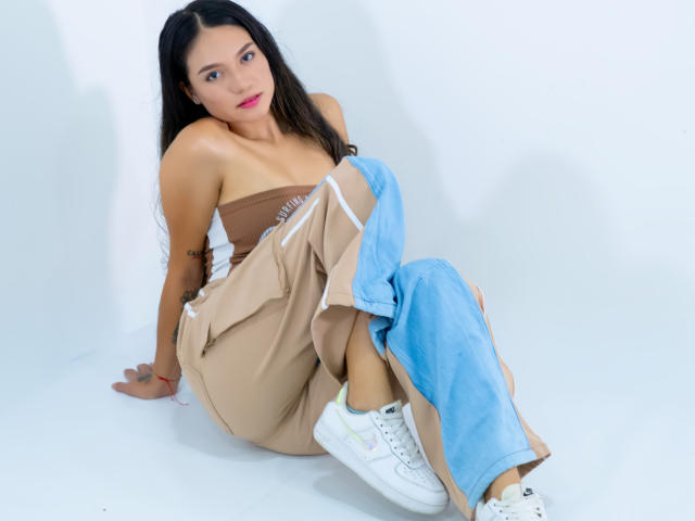 MillyPalmer with long black hair, brown strapless top, blue pants pulled down, showing white sneakers, tan pants, and small tits, sitting on white floor, looking at camera.